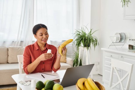 happy african american nutritionist holding apple and giving diet advice on laptop in kitchen