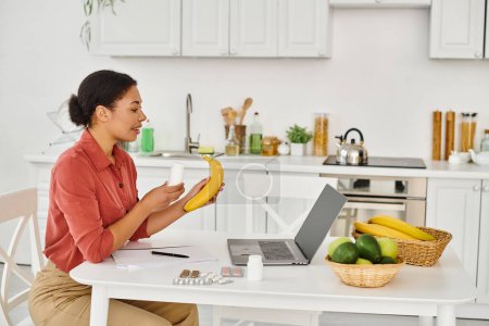 Téléchargez les photos : Heureux afro-américain nutritionniste tenant banane et donnant des conseils diététiques sur ordinateur portable dans la cuisine - en image libre de droit