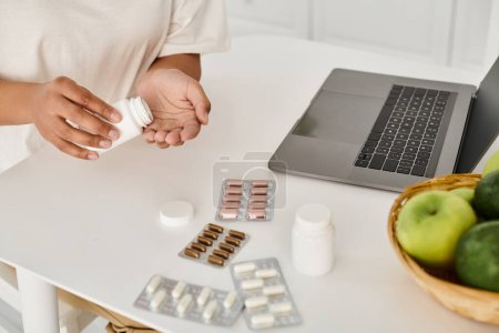 cropped african american woman holding bottle with pills near laptop during online consultation