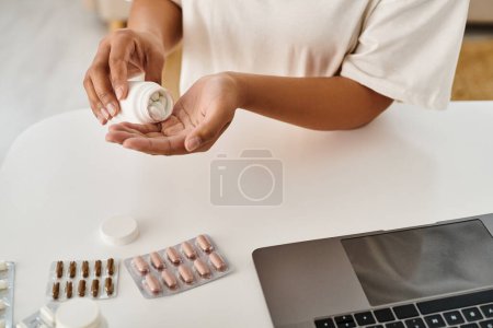 Téléchargez les photos : Cultivé afro-américain diététiste tenant bouteille avec des pilules près d'un ordinateur portable lors de la consultation en ligne - en image libre de droit