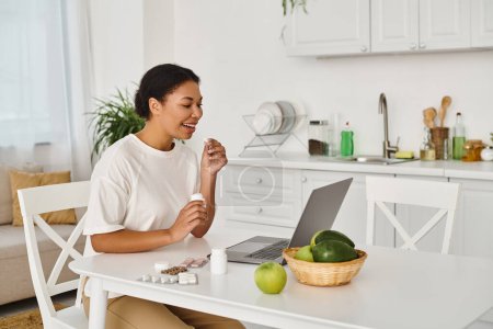 happy african american nutritionist taking medication in front of laptop near pills and fruits