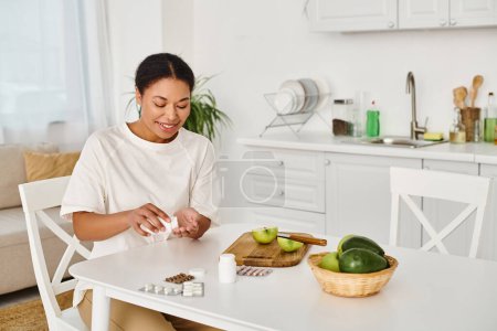 Téléchargez les photos : Heureux afro-américain nutritionniste compare les suppléments avec des fruits pour une alimentation saine à la maison - en image libre de droit