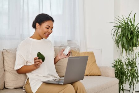 happy african american nutritionist with supplements and avocado giving dietary advice via laptop