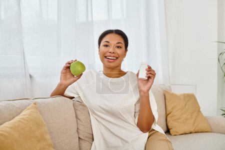 glücklich afrikanisch-amerikanische Ernährungsberaterin hält grünen Apfel und Nahrungsergänzungsmittel in der Hand und lächelt im Wohnzimmer
