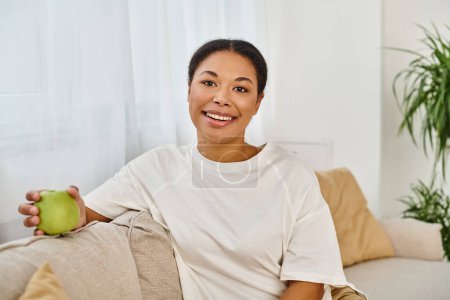 Radiant african american dietitian holding fresh green apple, relaxing on the living room sofa