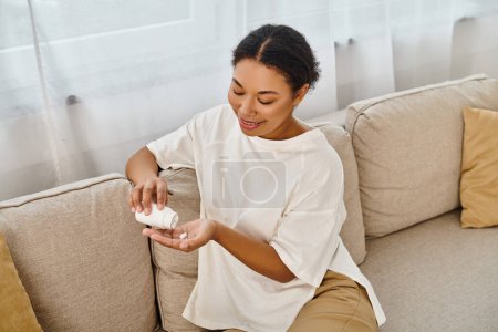 Téléchargez les photos : Heureuse diététiste afro-américaine verser des suppléments dans la main et se détendre sur le canapé dans le salon - en image libre de droit