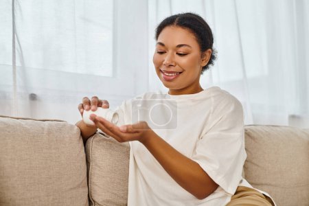 lächelnde afrikanisch-amerikanische Diätassistentin gießt Nahrungsergänzungsmittel in die Hand und entspannt sich auf dem Sofa im Wohnzimmer