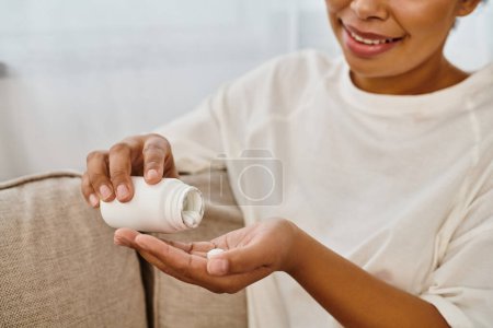 cropped happy african american woman pouring supplements into hand and relaxing on sofa at home