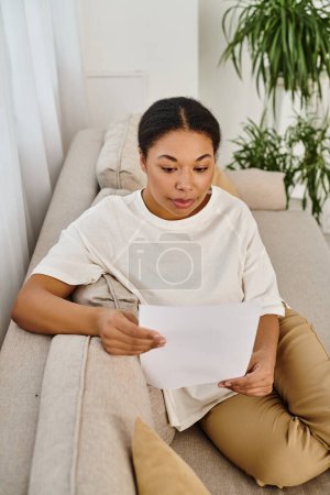 Photo for Young african american woman in casual attire reading dietary prescription and relaxing on sofa - Royalty Free Image