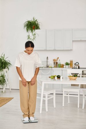 afrikanisch-amerikanische Frau in Freizeitkleidung, die in der Küche auf einer Waage steht, Gewichtsmanagement-Routine