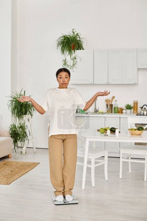 verwirrte schwarze Frau in Freizeitkleidung, die in der Küche auf einer Waage steht, Gewichtsmanagement-Routine