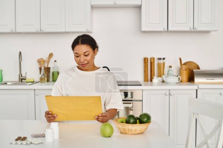 Photo for Brunette african american woman looking at envelope with dietary plan near supplements and fruits - Royalty Free Image