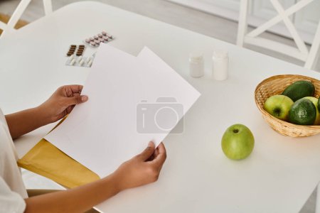 abgeschnittene Ansicht einer schwarzen Frau, die Ernährungsplan in der Nähe von Nahrungsergänzungsmitteln und Früchten auf dem Küchentisch überprüft
