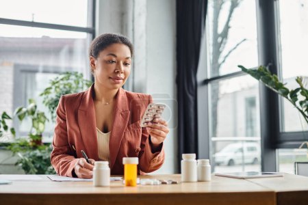 Photo for Curly african american nutritionist evaluating medication while writing notes in her office - Royalty Free Image