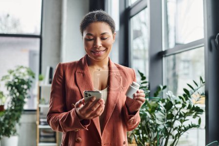 joyful african american nutritionist using smartphone while holding supplements in modern office