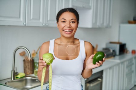mujer afroamericana alegre con cinta métrica y frutas que promueven una dieta saludable en la cocina