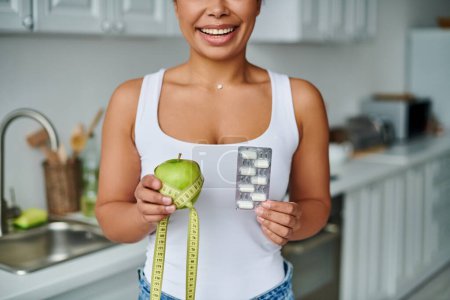 mujer afroamericana feliz recortada con cinta métrica con manzana y paquete con suplementos