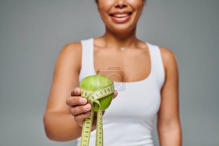 cropped view of happy african american woman with measuring tape and apple on grey background