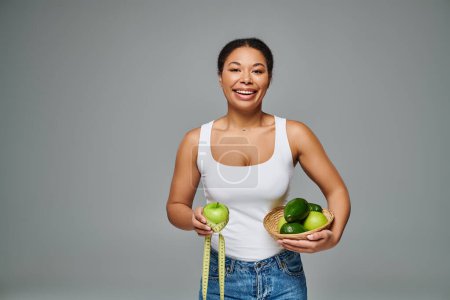 heureuse diététiste afro-américaine avec pomme et suppléments mettant en valeur la santé sur fond gris