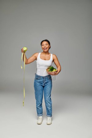 cheerful african american dietitian with apple and supplements showcasing health on grey backdrop