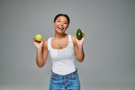 Mujer afroamericana radiante con manzana y aguacate promoviendo una nutrición equilibrada sobre fondo gris