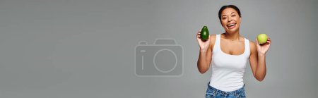 Mujer afroamericana radiante con manzana y aguacate promoviendo una nutrición equilibrada en gris, estandarte