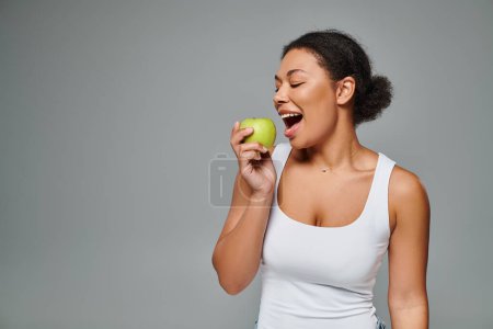 mujer afroamericana feliz con dientes blancos mordiendo manzana verde sobre fondo gris, alimentación saludable