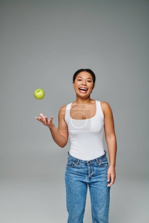 Photo for Happy african american woman with white teeth throwing green apple in air on grey background - Royalty Free Image