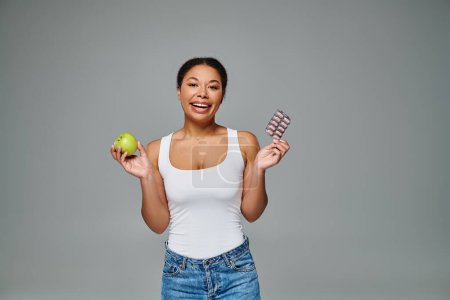 glücklich afrikanisch-amerikanische Frau vergleicht Nahrungsergänzungsmittel mit grünem Apfelgrauen Hintergrund, Diätwahl