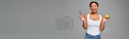 happy african american woman comparing supplements with green apple grey background, banner