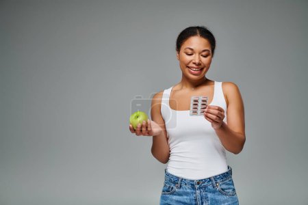 happy african american woman comparing supplements with green apple grey background, balanced diet