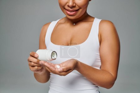 femme afro-américaine cultivée versant des pilules de la bouteille dans la main sur fond gris, plan alimentaire