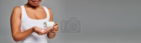 pancarta de la mujer afroamericana recortada vertiendo píldoras de la botella en la mano sobre fondo gris
