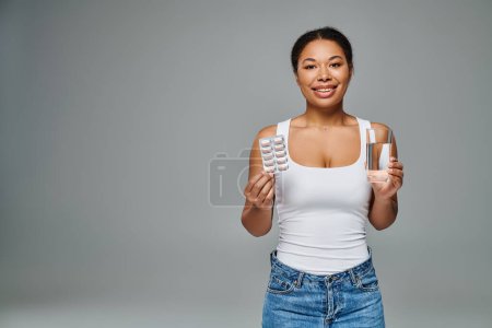 Femme afro-américaine souriante tenant des vitamines dans une plaquette thermoformée et un verre d'eau sur gris