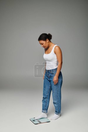 fröhliche junge afrikanisch-amerikanische Frau in lässiger Kleidung, die auf einer Waage steht, grauer Hintergrund