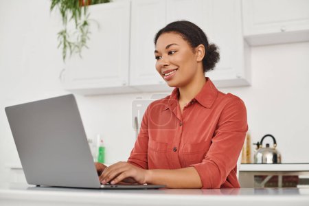 Téléchargez les photos : Souriant frisé afro-américain diététiste en utilisant son ordinateur portable tout en tapant et en travaillant à la maison - en image libre de droit