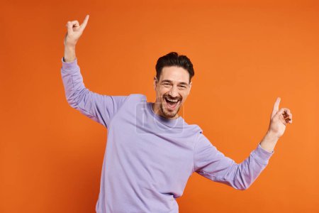 joyful and bearded man in purple sweatshirt dancing with hands up on vibrant orange background