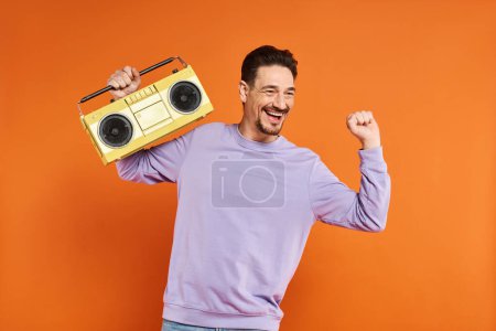 fröhlicher und bärtiger Mann in lila Sweatshirt mit Retro-Boombox auf orangefarbenem Hintergrund, Musik