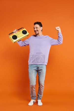 aufgeregter und bärtiger Mann in lila Sweatshirt mit Retro-Boombox auf orangefarbenem Hintergrund, Musik