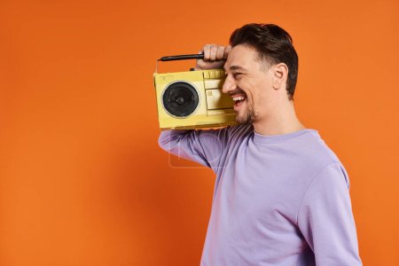 fröhlicher Mann in lila Sweatshirt mit Retro-Boombox und Musik auf orangefarbenem Hintergrund