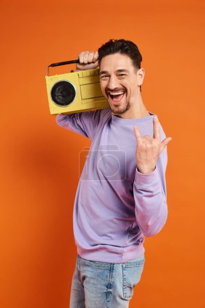 excited and bearded man showing rock sign and holding retro boombox on orange background, music