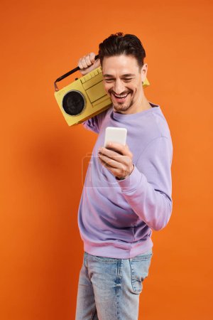 joyful and bearded man using smartphone and holding retro boombox on orange background, music