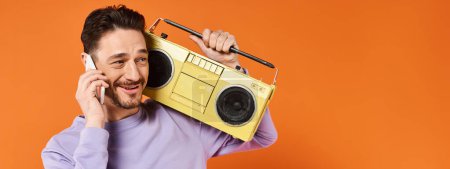 joyful and bearded man talking on smartphone and holding retro boombox on orange background, banner
