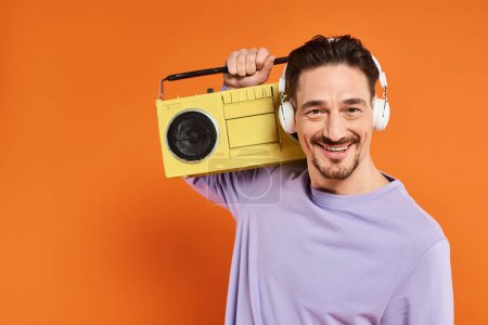 happy bearded man in purple sweater and wireless headphones holding boombox on orange background