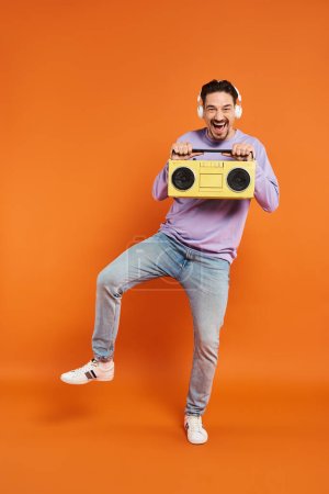 fröhlicher bärtiger Mann in lila Pullover und kabellosen Kopfhörern mit Boombox auf orangefarbenem Hintergrund