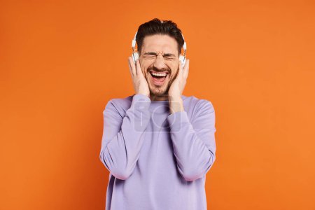 excited bearded man in purple sweater and wireless headphones listening music on orange background
