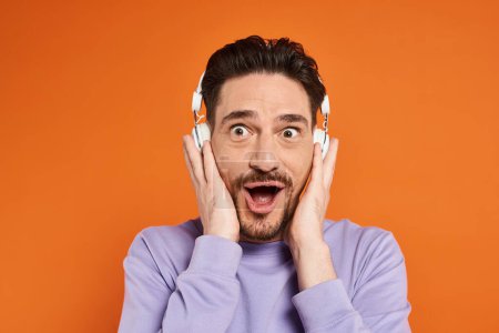 amazed bearded man in purple sweater and wireless headphones listening music on orange background