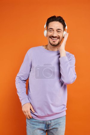 glücklicher bärtiger Mann in lila Pullover und drahtlosen Kopfhörern, der Musik auf orangefarbenem Hintergrund hört