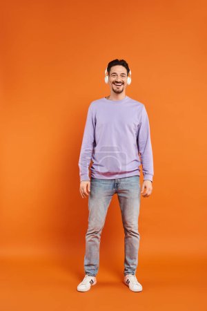 cheerful bearded man in purple sweater and wireless headphones listening music on orange background