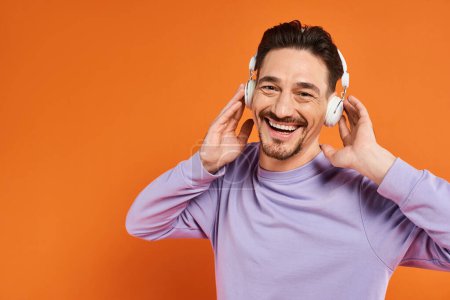 positiver Mann in lila Pullover und drahtlosen Kopfhörern, der Musik auf orangefarbenem Hintergrund hört